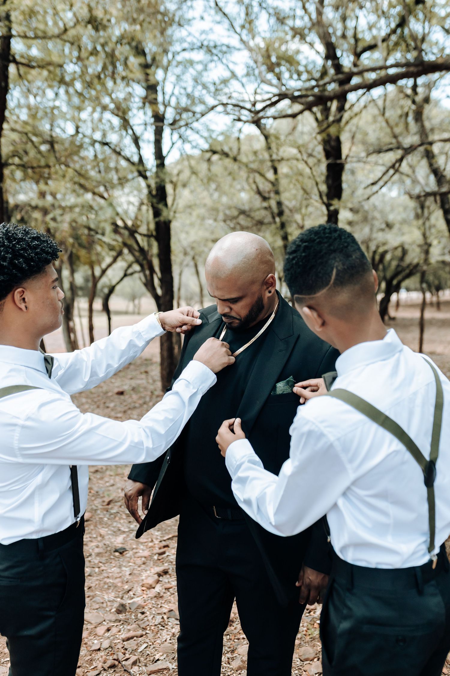 Two people in white shirts adjusting the tie of person in black suit outdoors.