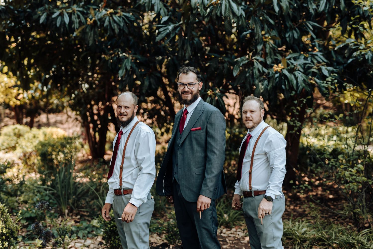Three groomsmen pose casually in outdoor setting with lush greenery background.