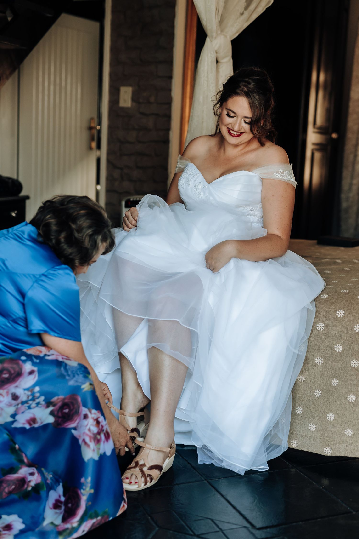 Bride in a flowing white dress gets help putting on her wedding shoes.