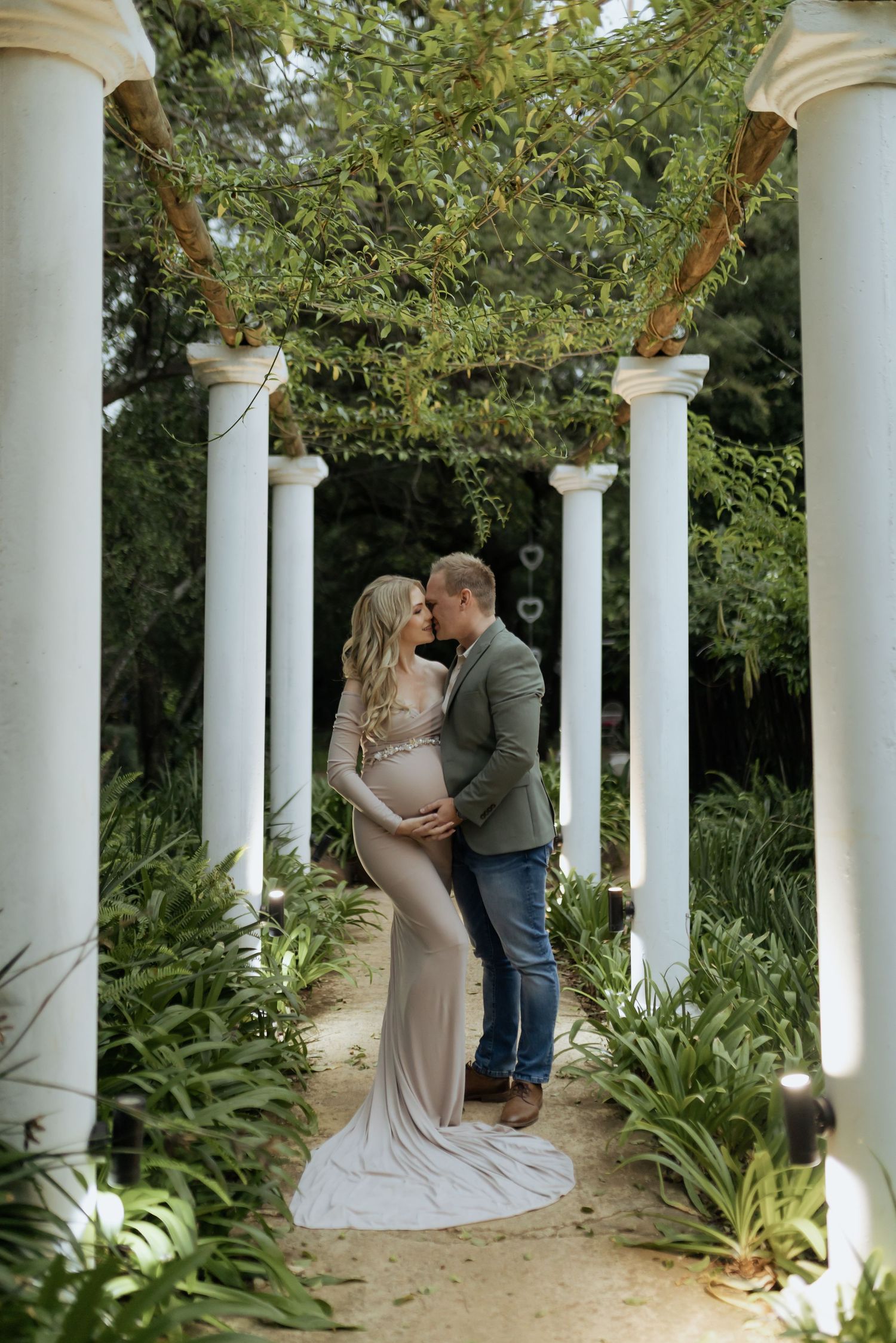 A romantic moment captured between a couple in a white columned garden walkway surrounded by lush greenery.