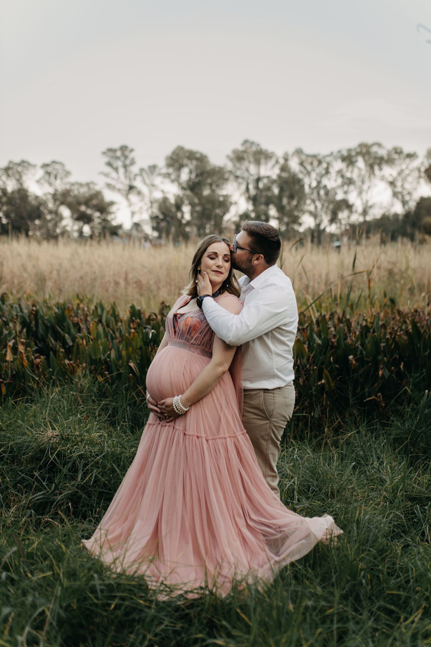 A romantic couple embraces in a field of tall grass, with her wearing a flowing pink dress during a sunset photoshoot.