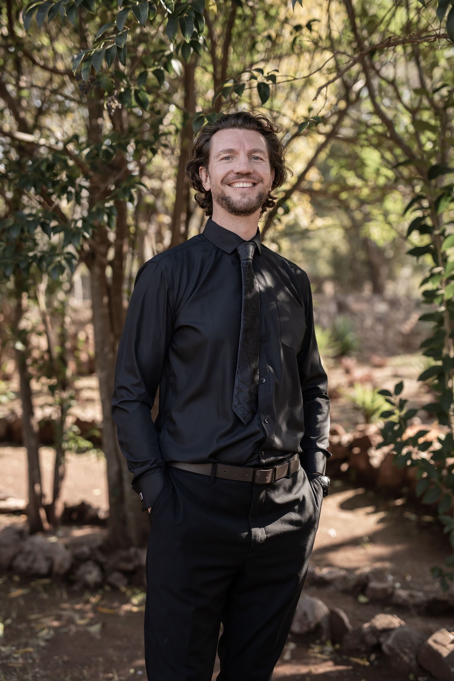 A person in all black attire smiles warmly while standing in a wooded outdoor setting with dappled sunlight.