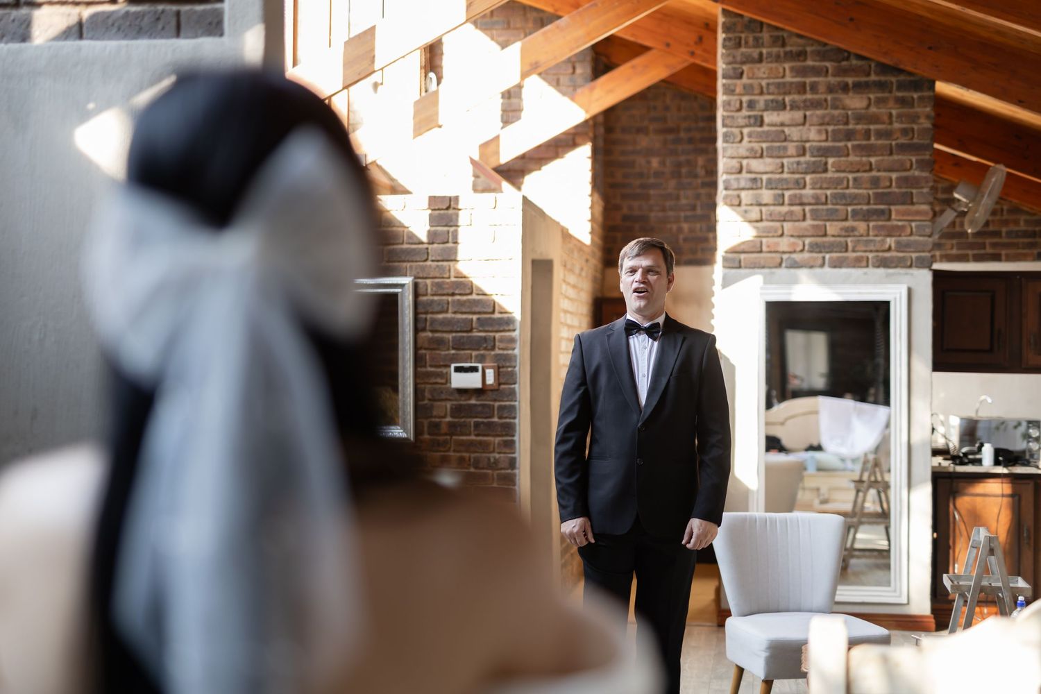 A well-dressed person in a black tuxedo stands in front of an elegant brick interior with wood beam accents.