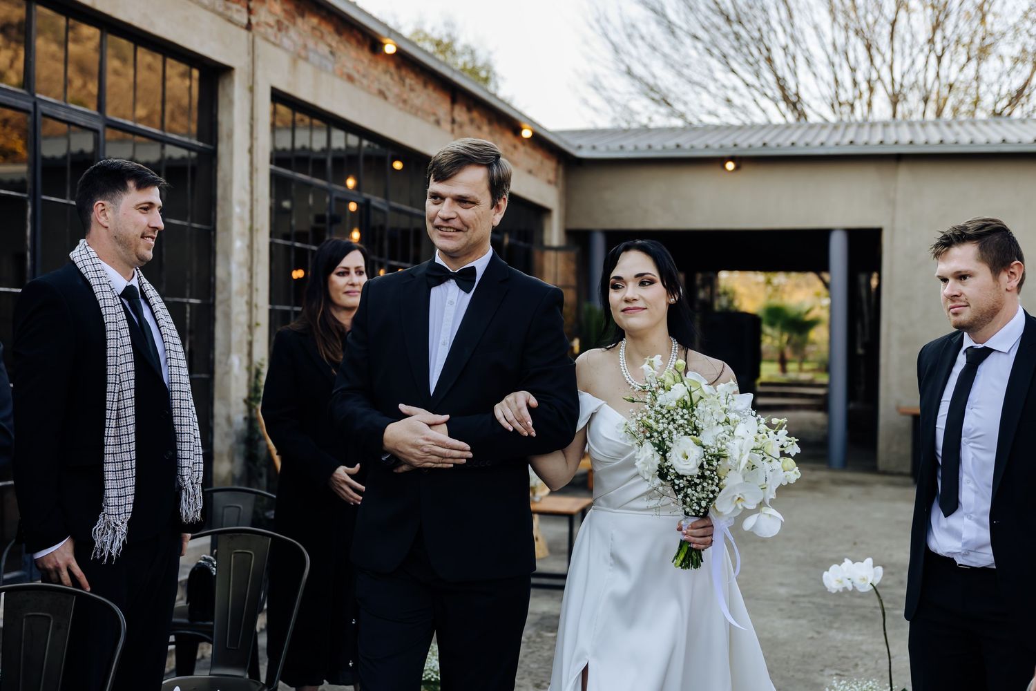 A wedding party laughs together while walking outside a rustic venue building on a winter day.