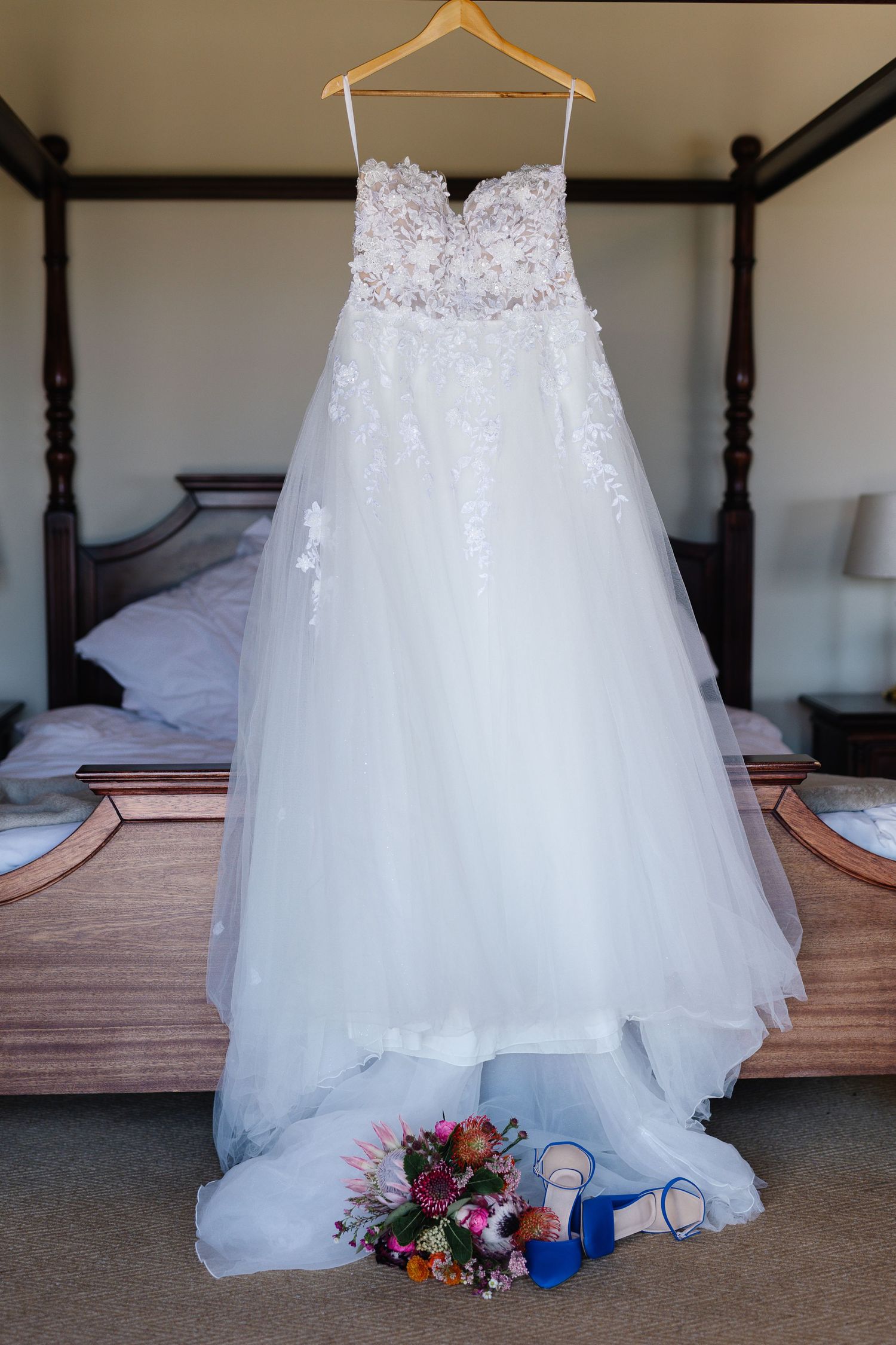 A white lace wedding dress hangs on a wooden hanger above a bed with a colorful floral bouquet below.