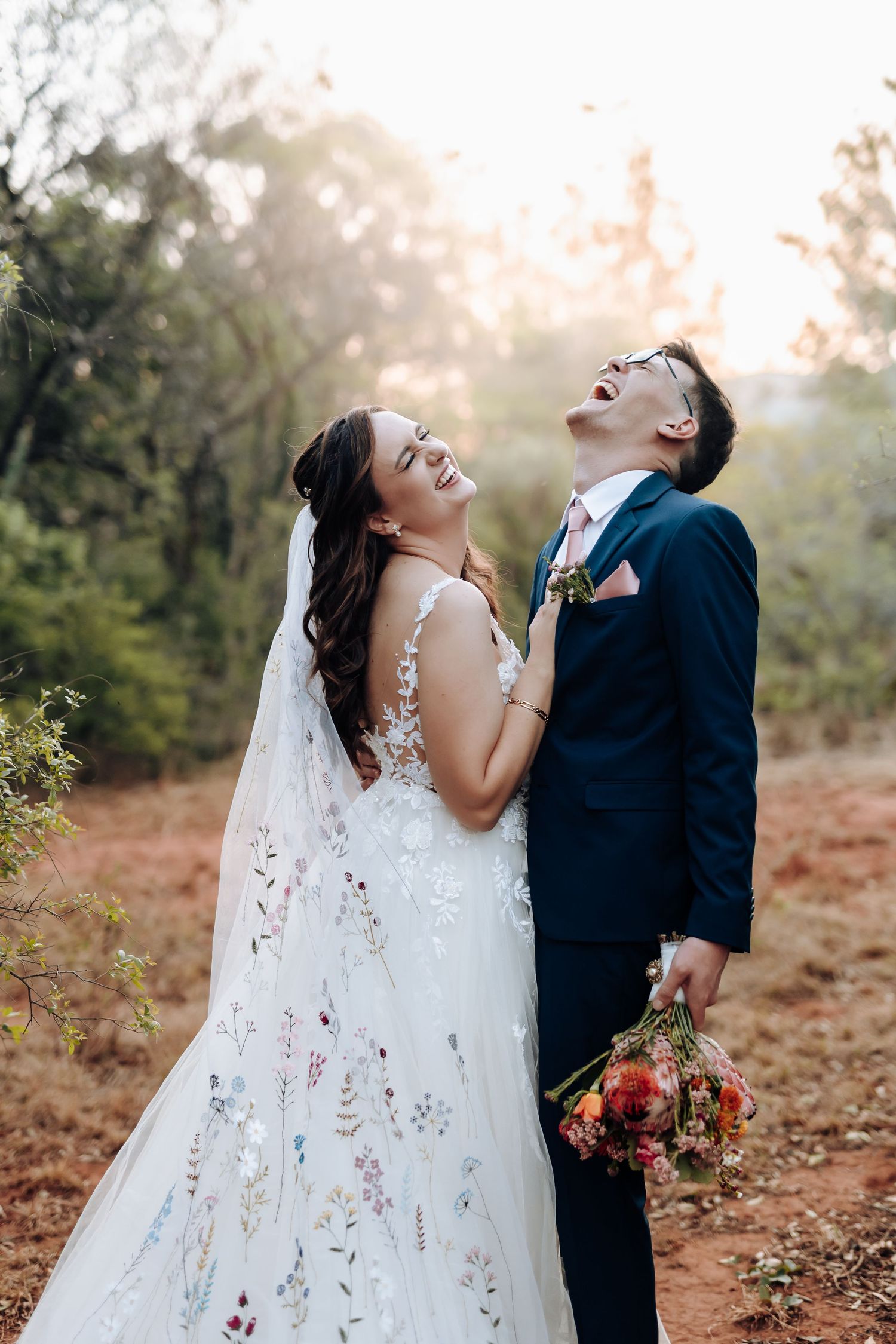 A happy couple shares joyful laughter together during their autumn wedding photoshoot in a wooded setting.