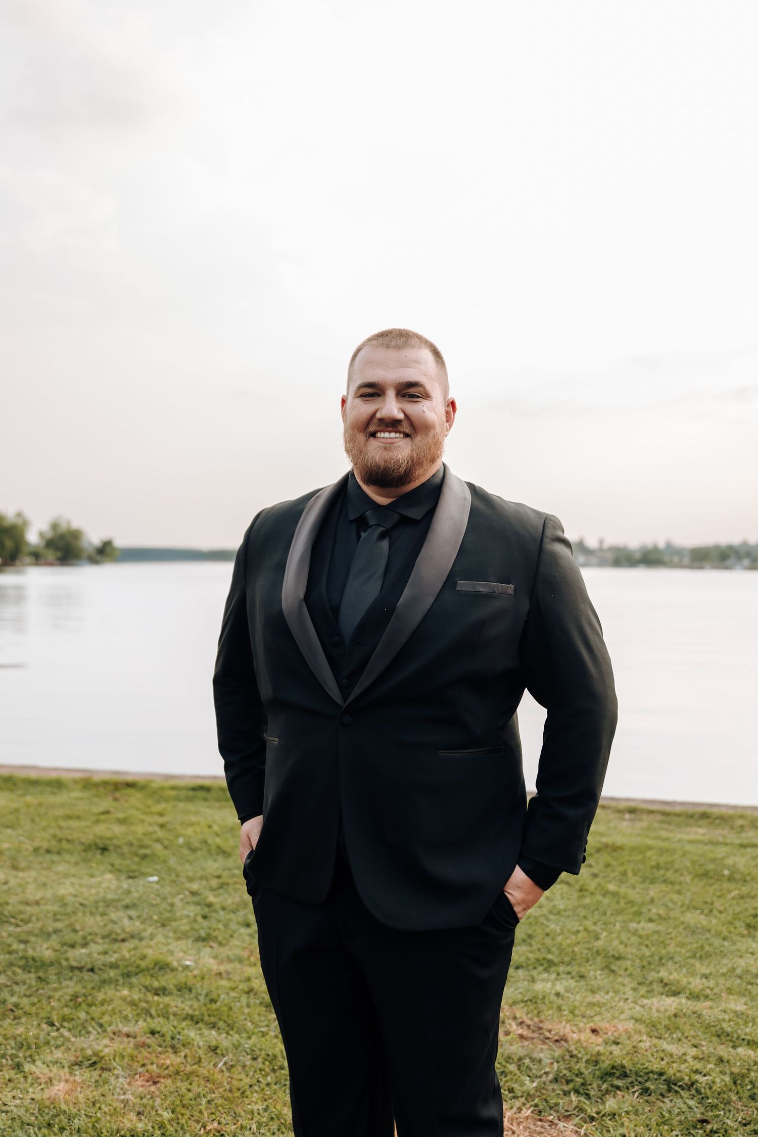 A person in a black suit smiles while standing outdoors near a waterfront on a grassy area.