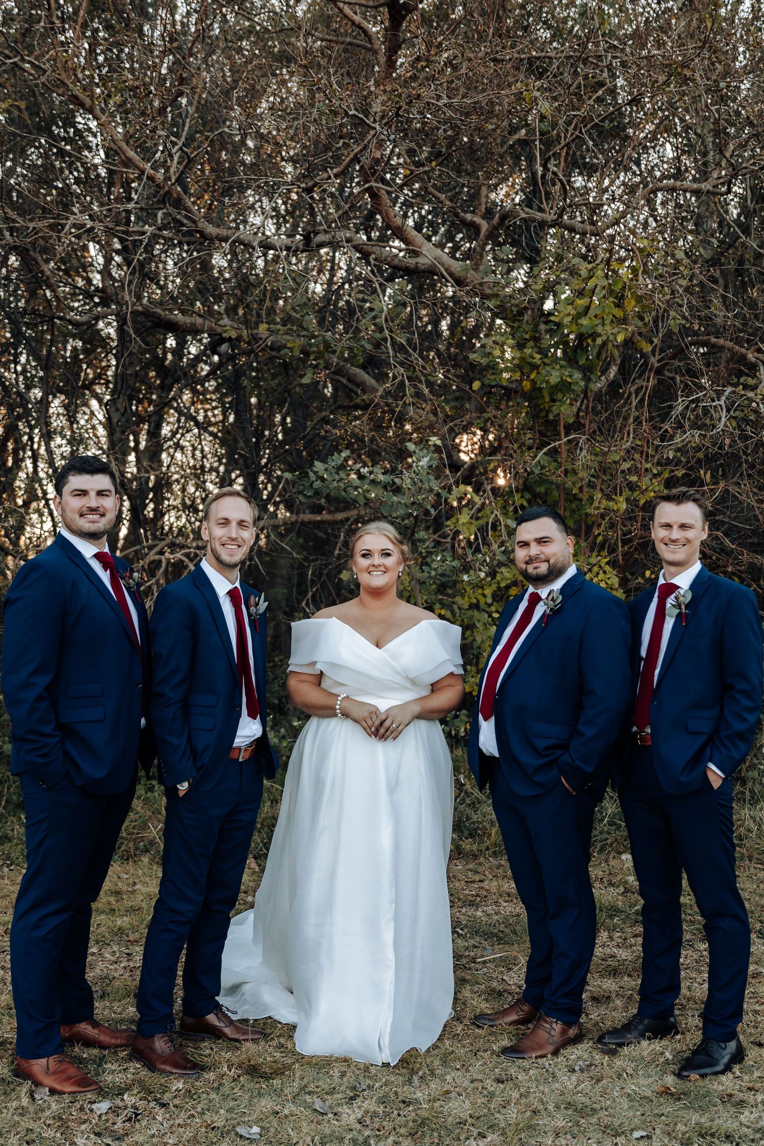 Bride in white off-shoulder gown poses with four groomsmen in navy suits and red ties outdoors.