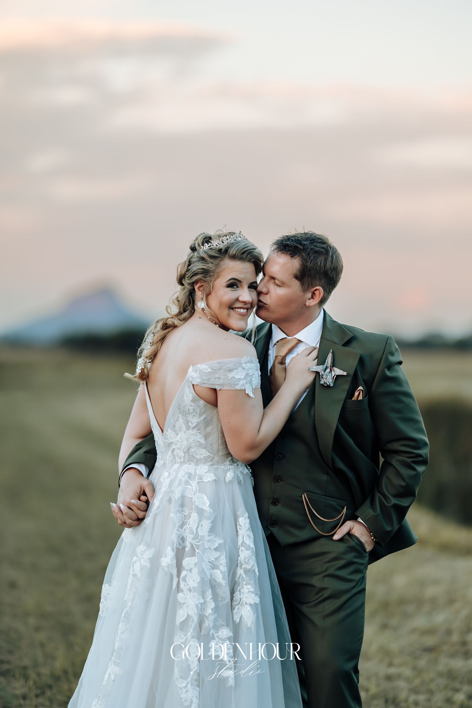 Bride in lace off-shoulder gown and groom in green suit share a tender moment at sunset outdoors.