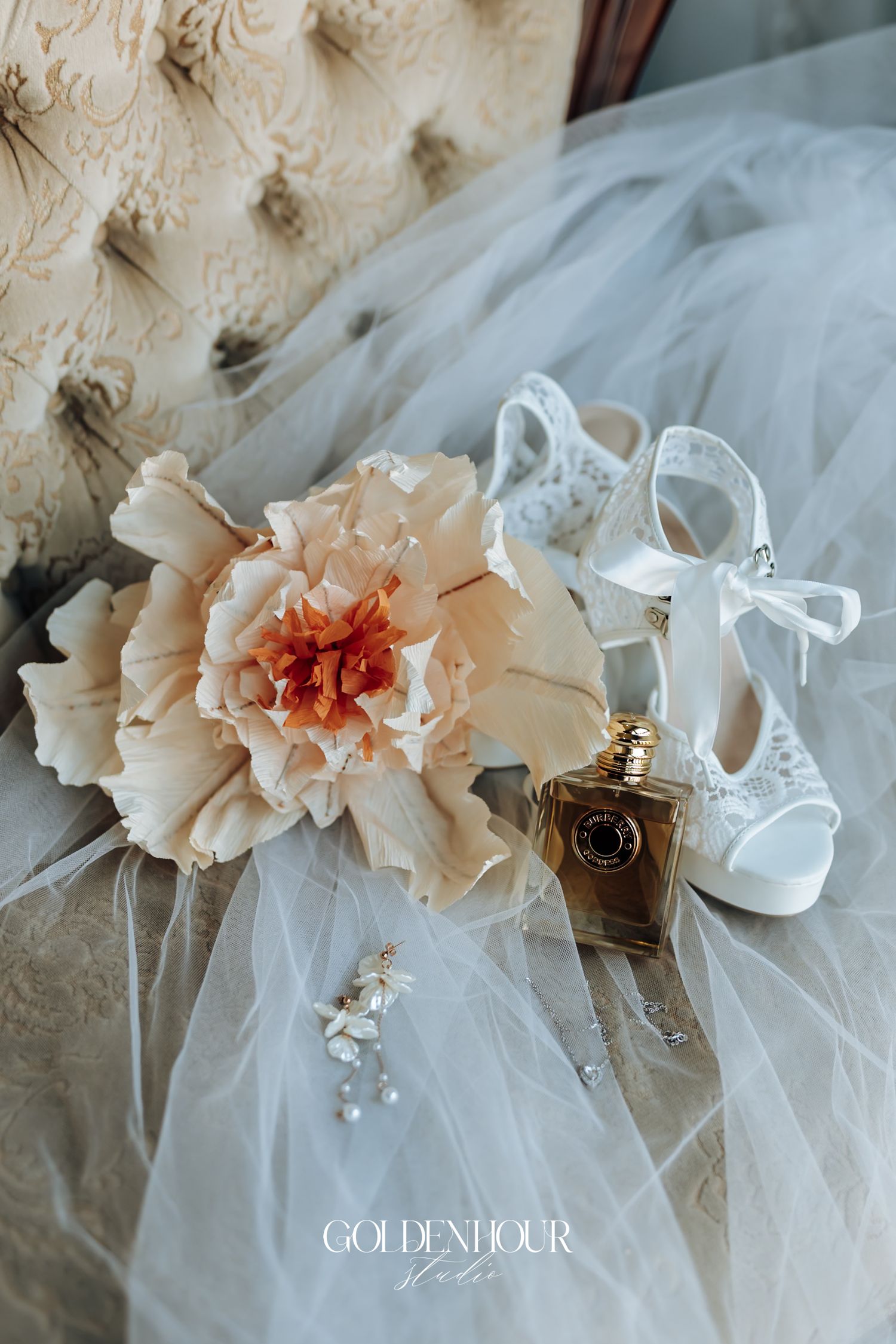 Bridal flat lay with a peach peony, white lace heels, gold perfume bottle, and delicate earrings on a tulle veil.
