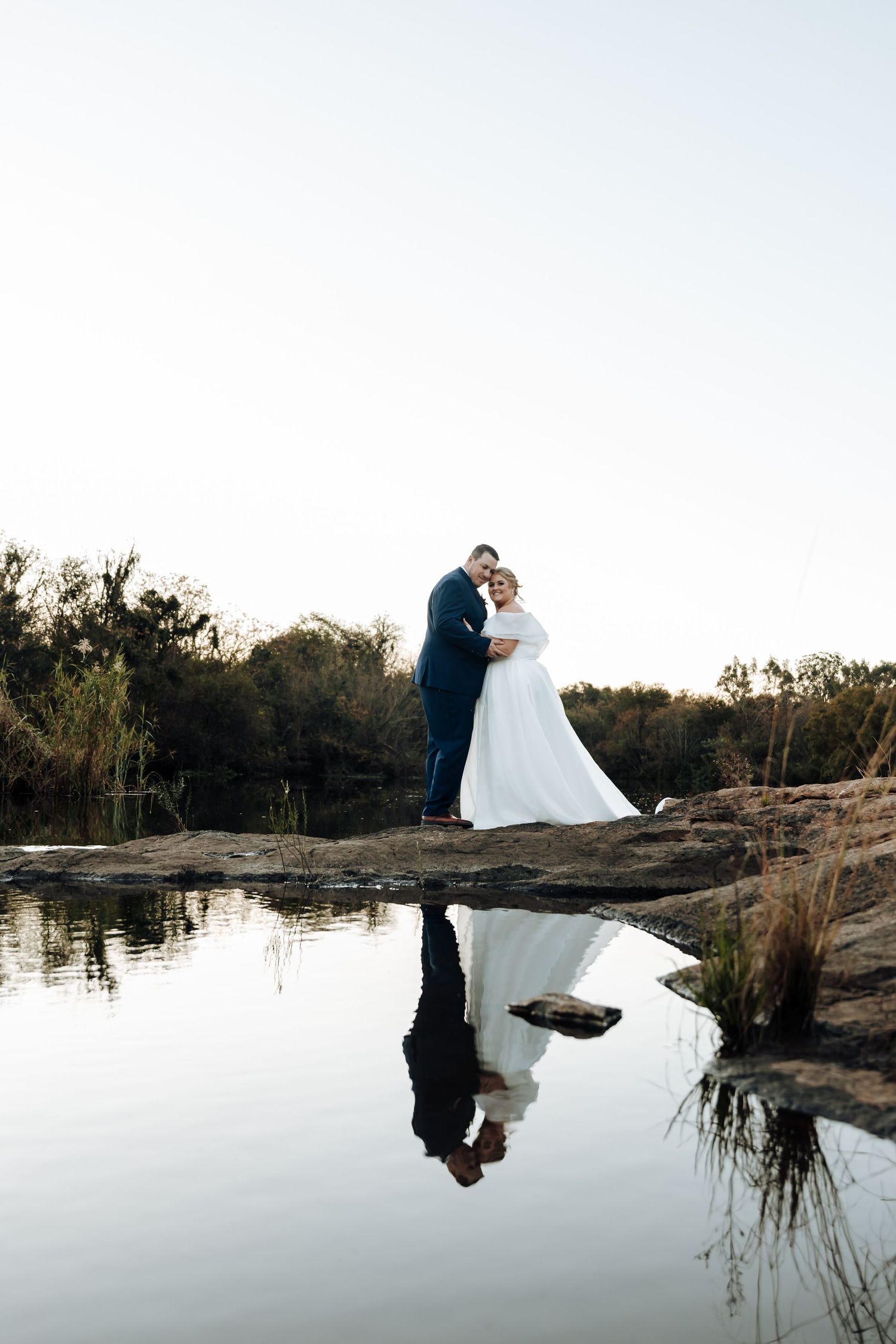 Bride and groom embracing on rocky waterfront with their reflection mirrored in the still water at golden hour.