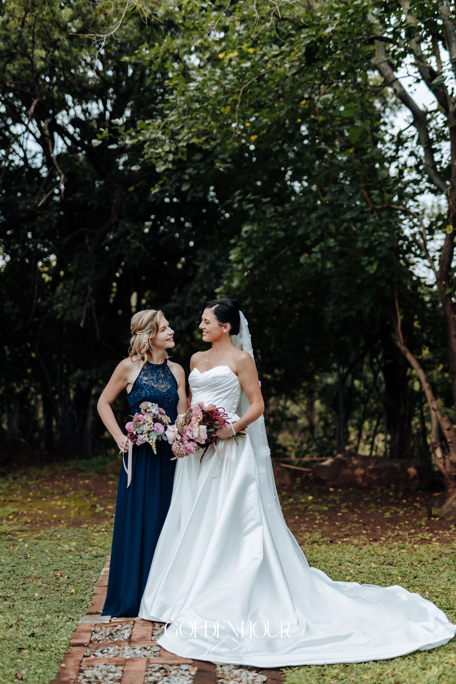 Bride in a white satin gown with train stands beside bridesmaid in navy dress in lush garden setting.