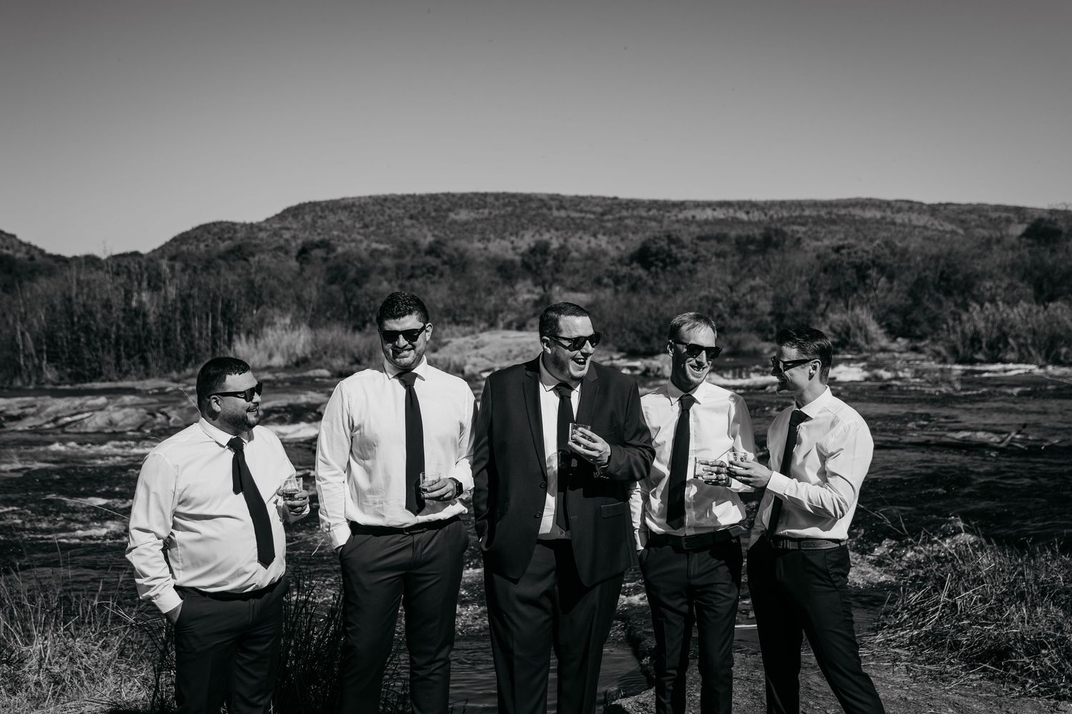 Five groomsmen in black ties and sunglasses pose outdoors in a rocky landscape for a wedding photo.