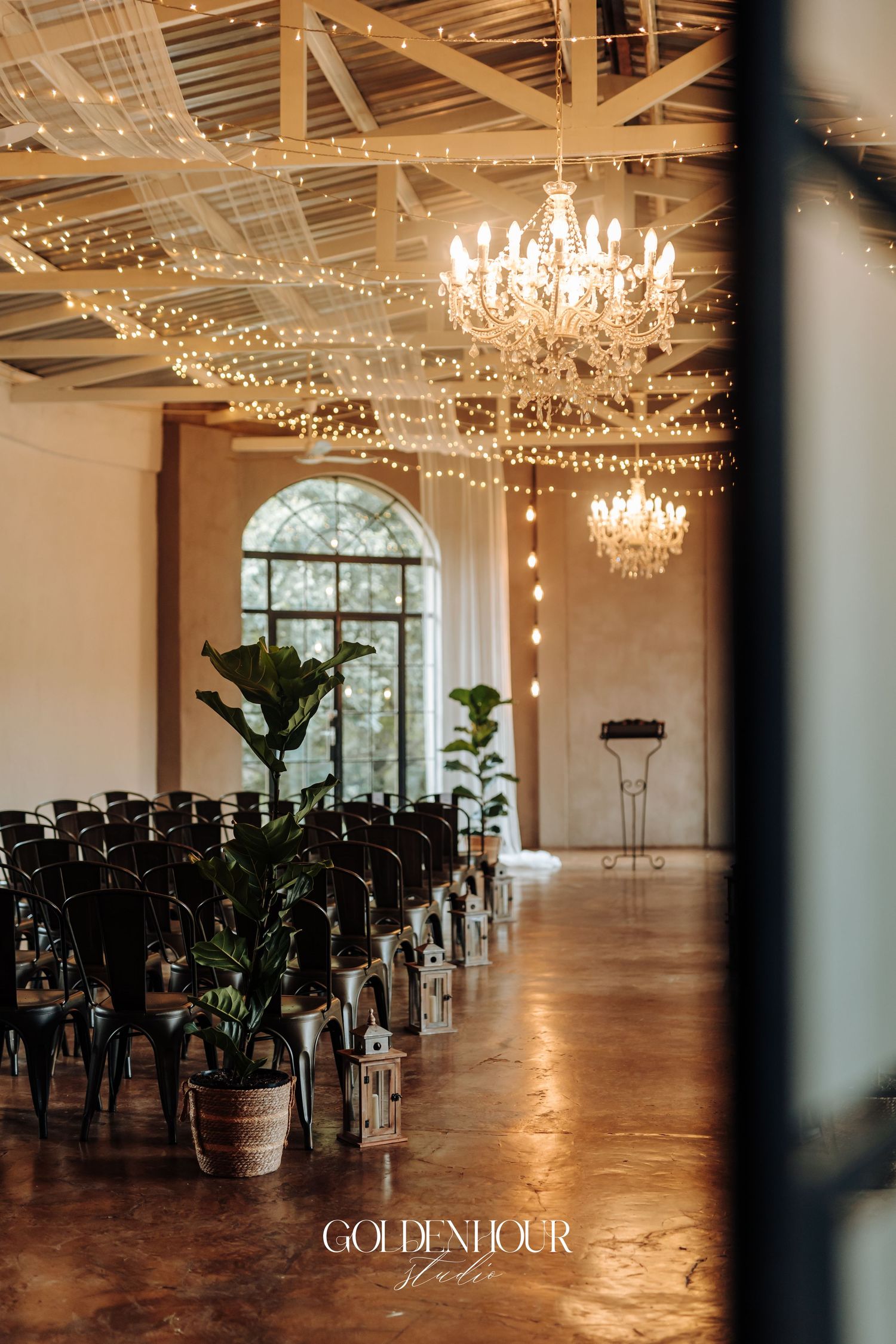 Elegant wedding ceremony venue with fairy lights, crystal chandeliers, black chairs, and potted plants in a bright hall.