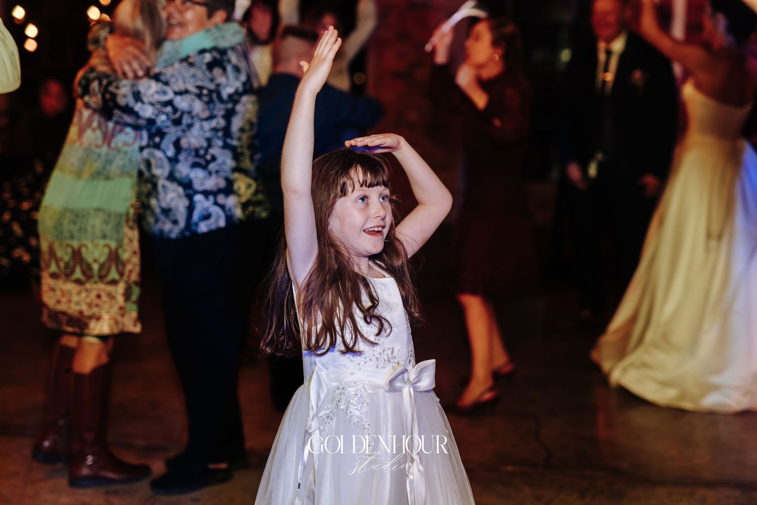 Young flower girl in white dress dances joyfully among guests on a wedding reception dance floor.