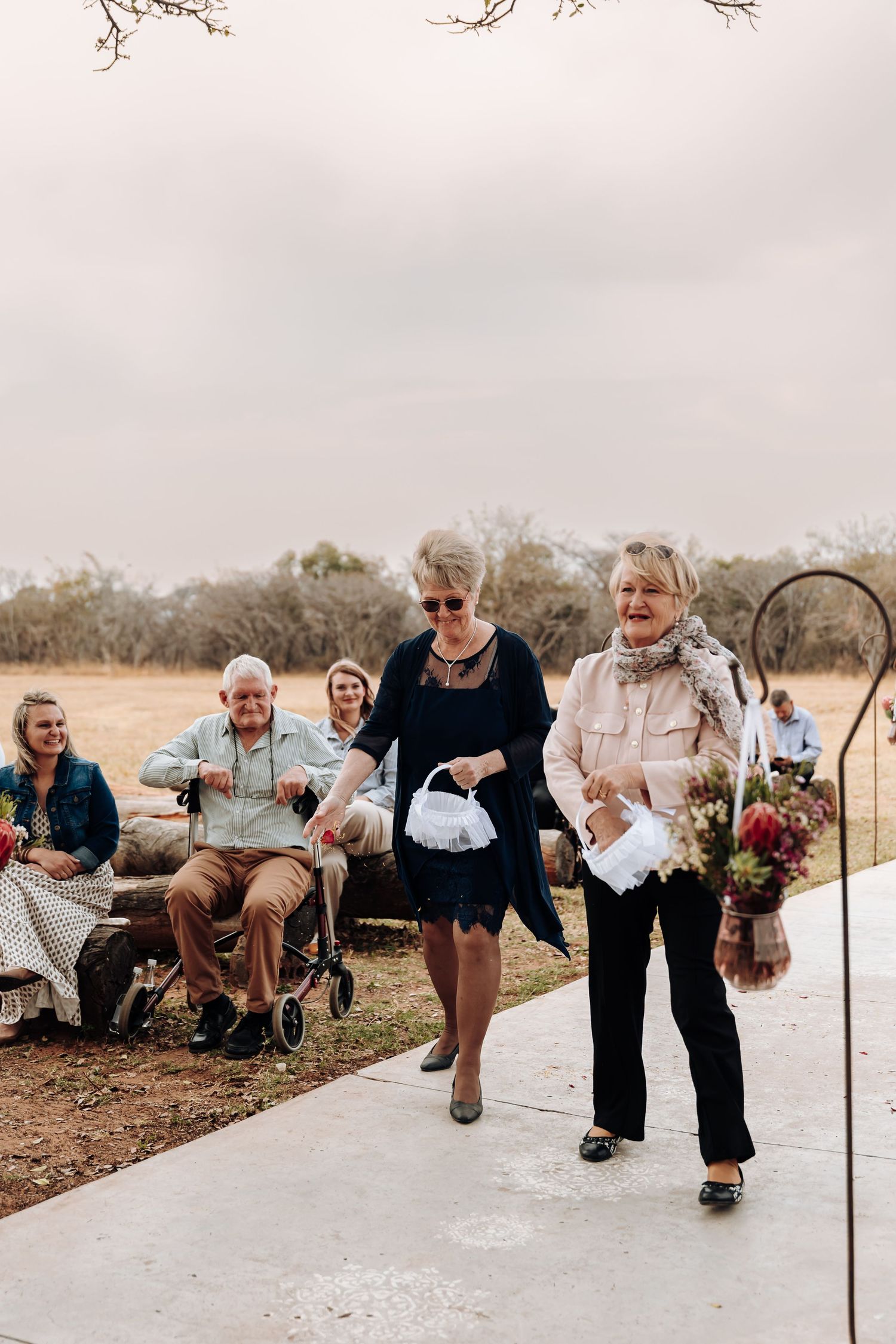 Wedding guests walk down an outdoor path while carrying flowers at a rustic outdoor ceremony venue.