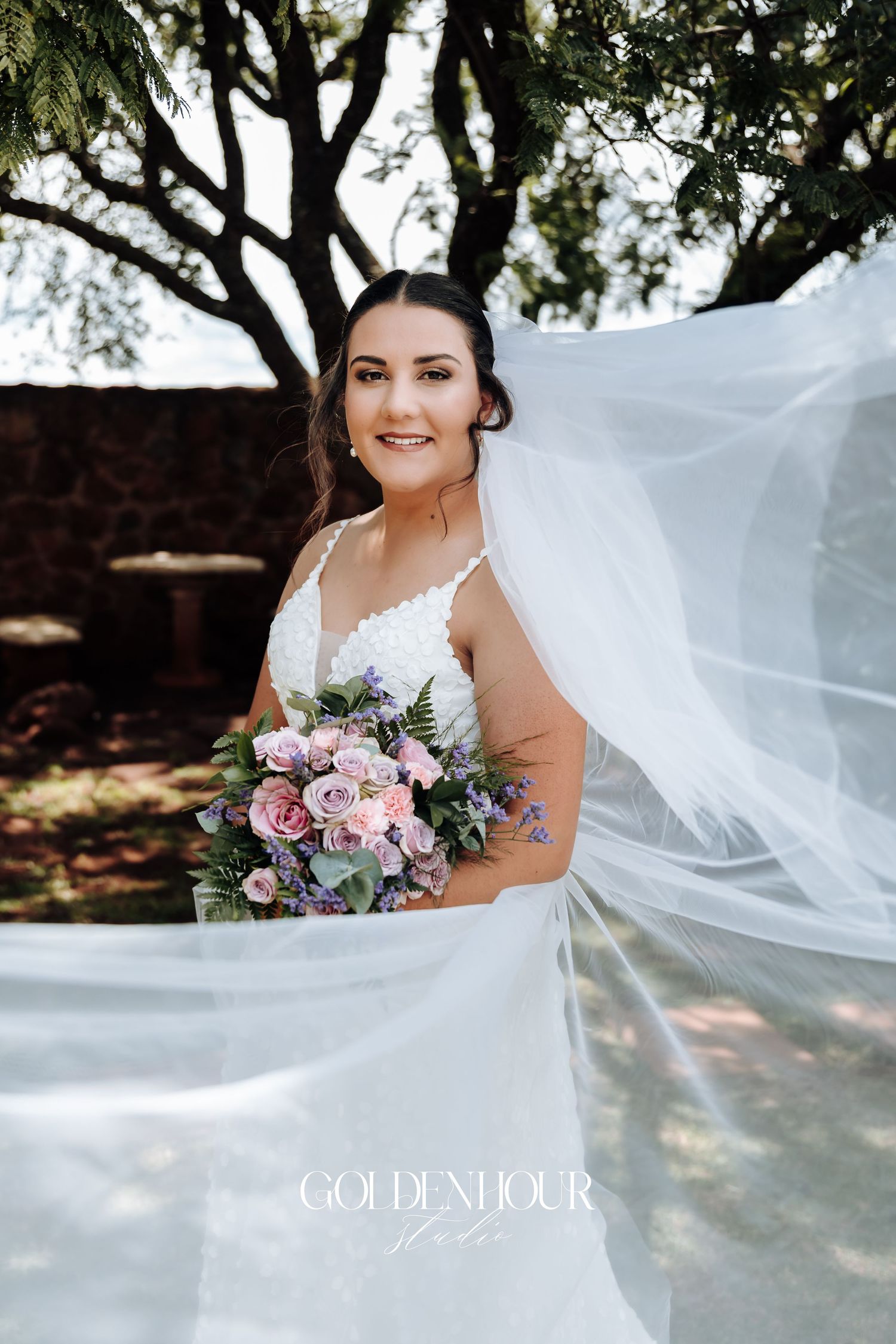 A flowing veil catches the breeze as person holds pink and purple bouquet in garden setting.