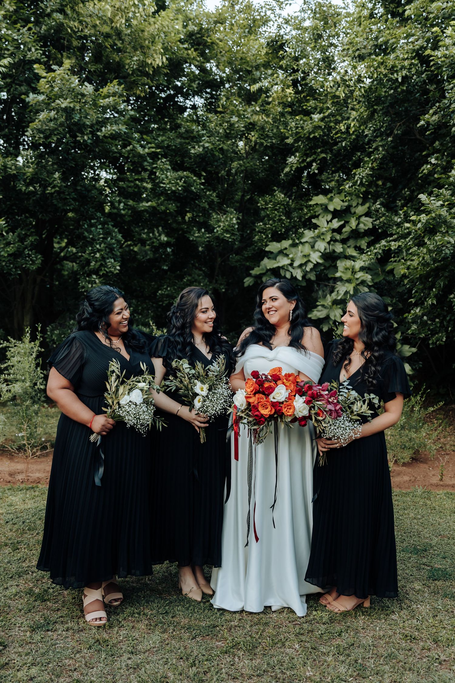 Bridal party in black dresses sits together on grass during outdoor photo session.