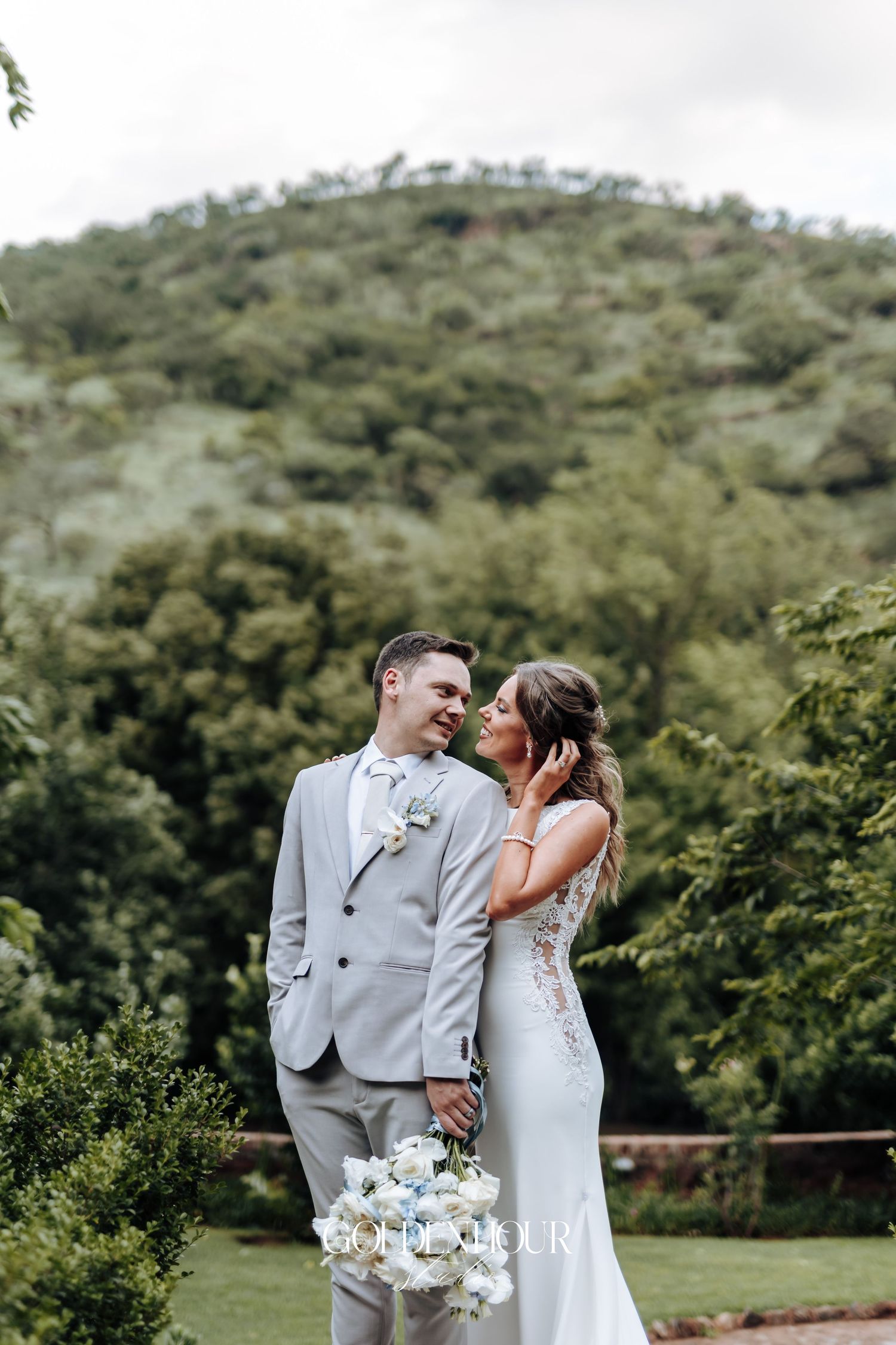 Newlyweds share an intimate moment in a lush green outdoor setting with mountains in the background.