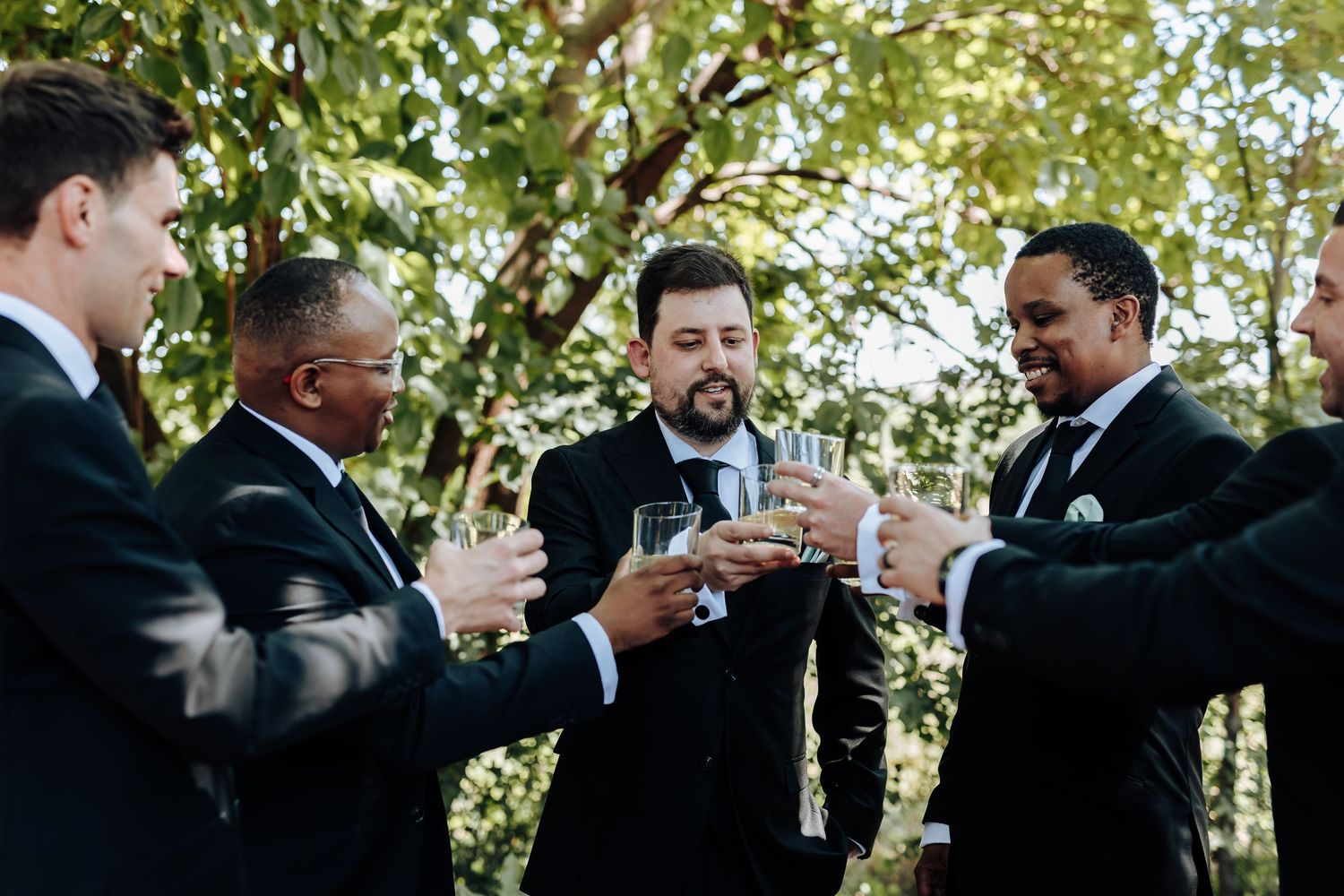 A group in formal black suits toasting with champagne glasses outdoors under green trees.