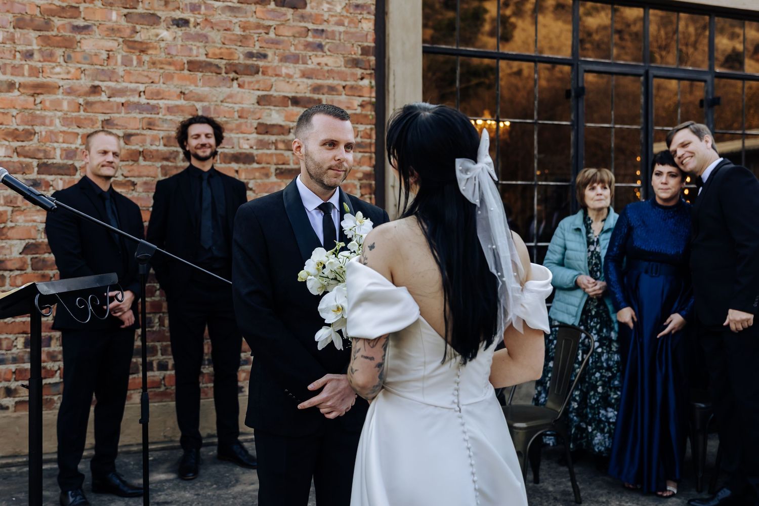 Wedding ceremony moment outside rustic brick building with couple exchanging vows while guests look on.