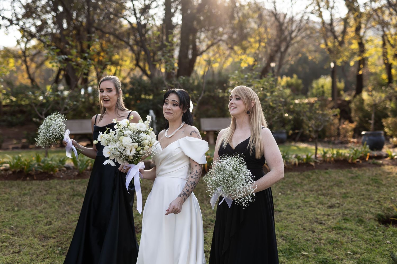 Three people in formal attire hold white floral bouquets while standing outdoors in a sunlit garden setting.