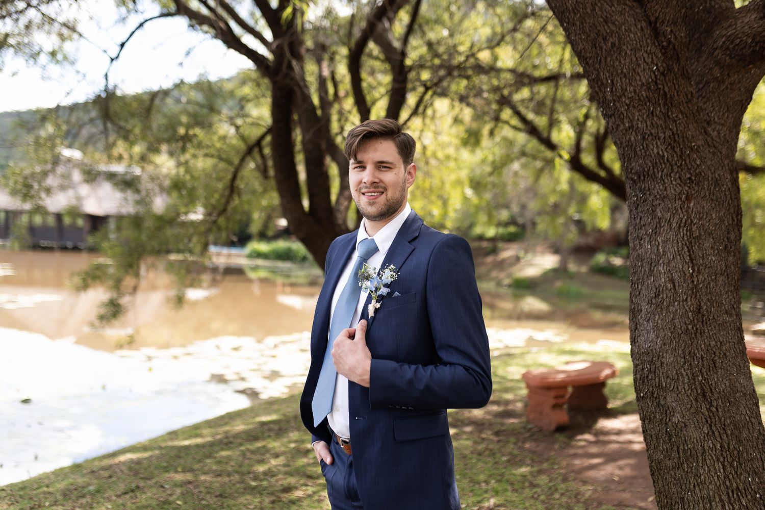 Groom in classic navy suit poses against natural lake backdrop.