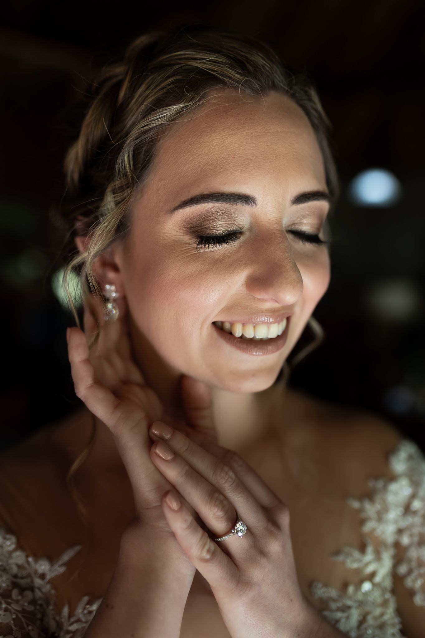 A closeup portrait of a bride wearing a lace gown and engagement ring, smiling with closed eyes in soft lighting.