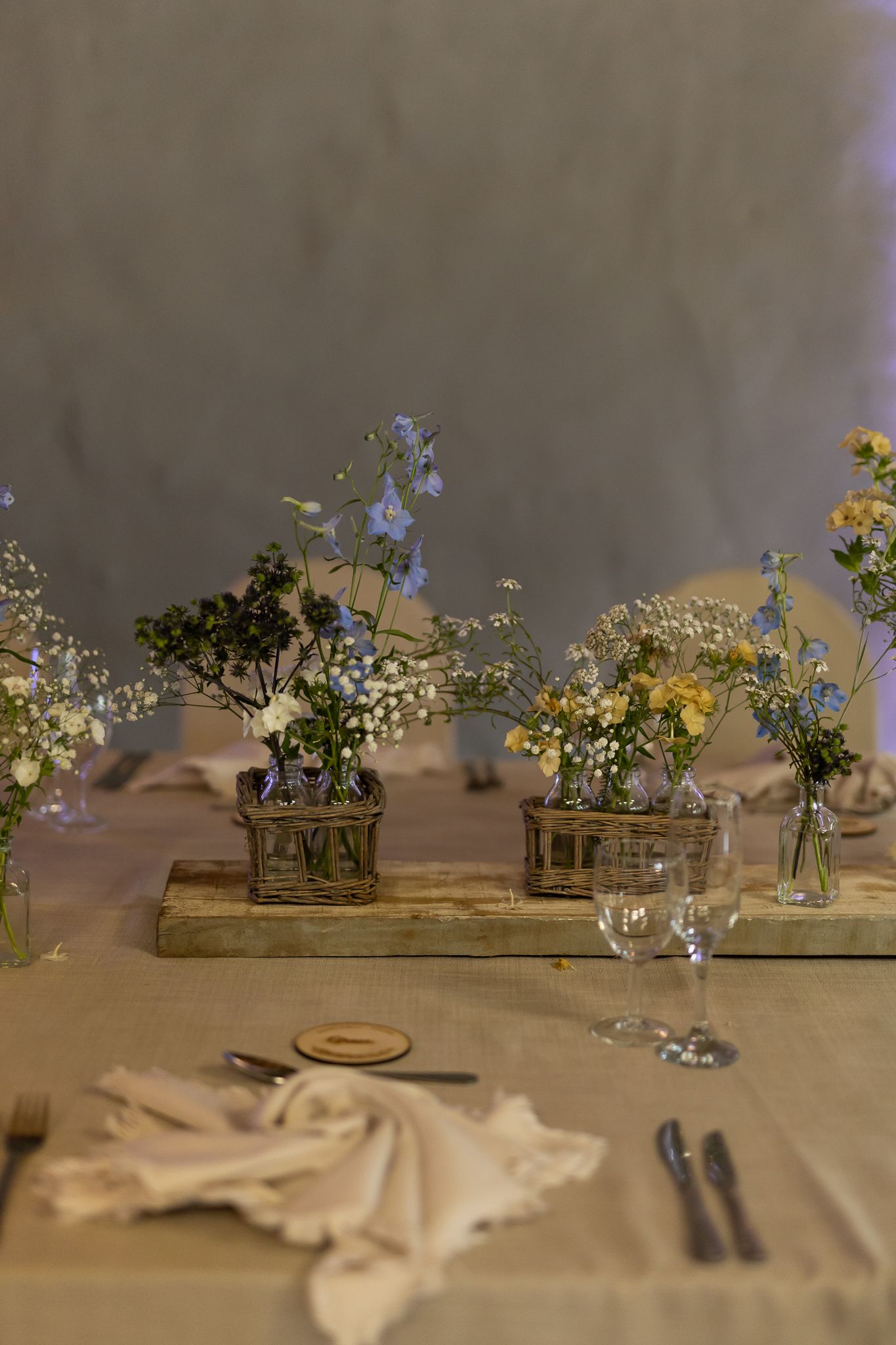 Rustic wedding table decor with wildflowers in glass vases arranged on wooden plank.