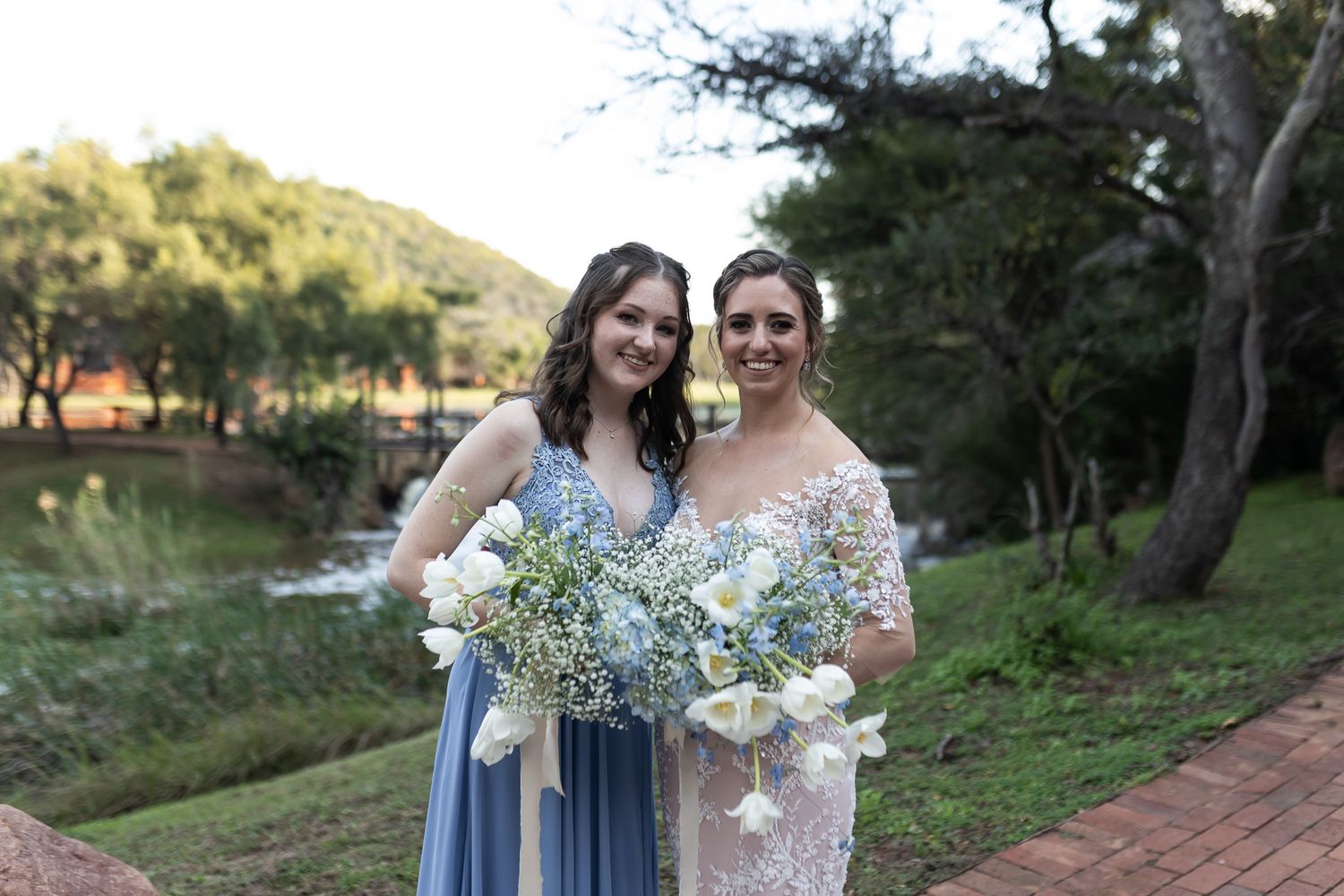Two attendants hold white and green bouquets while posing by lakeside during wedding.