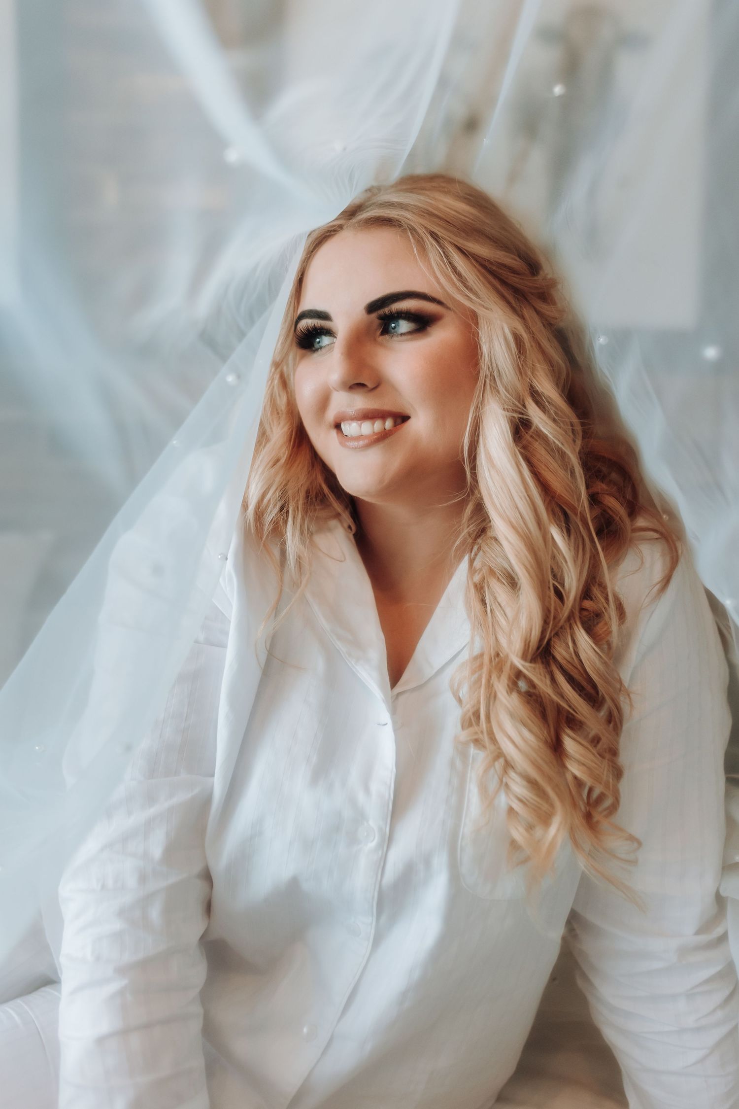 A portrait with flowing blonde waves and glamorous makeup against a soft white veil backdrop.