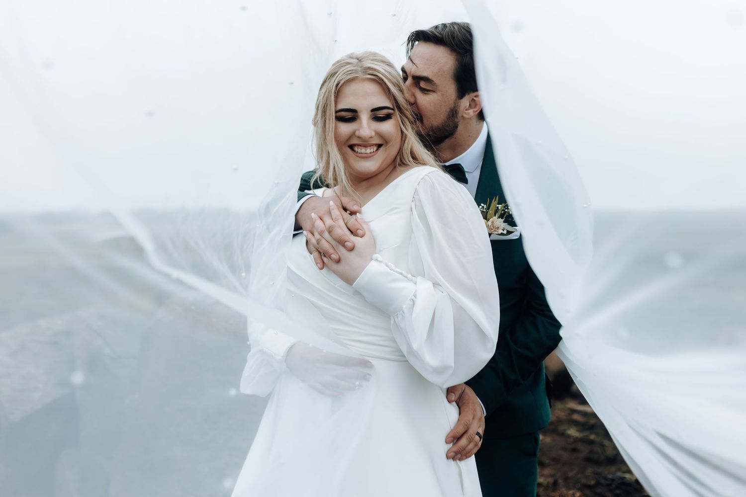Joyful wedding couple laughs together while embracing outdoors with flowing white veil and dress.