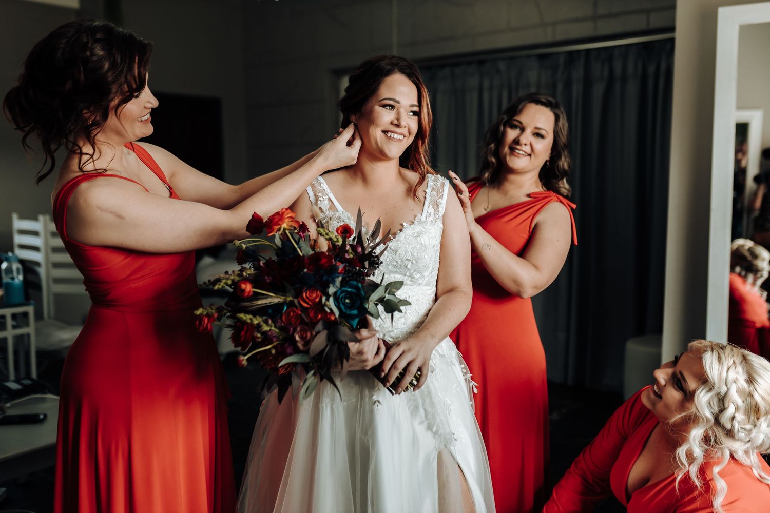 Bridesmaids in red dresses help style the bride's hair while she holds a dark floral bouquet.