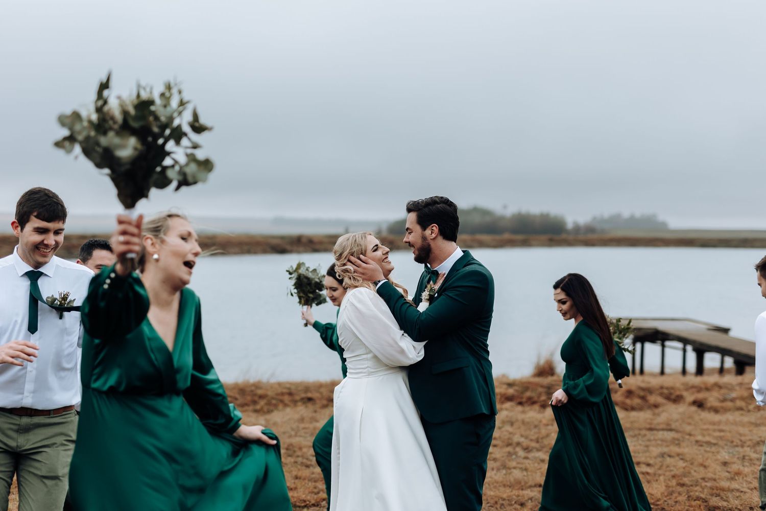 Wedding party in emerald green dresses gather by a waterfront on an overcast day.