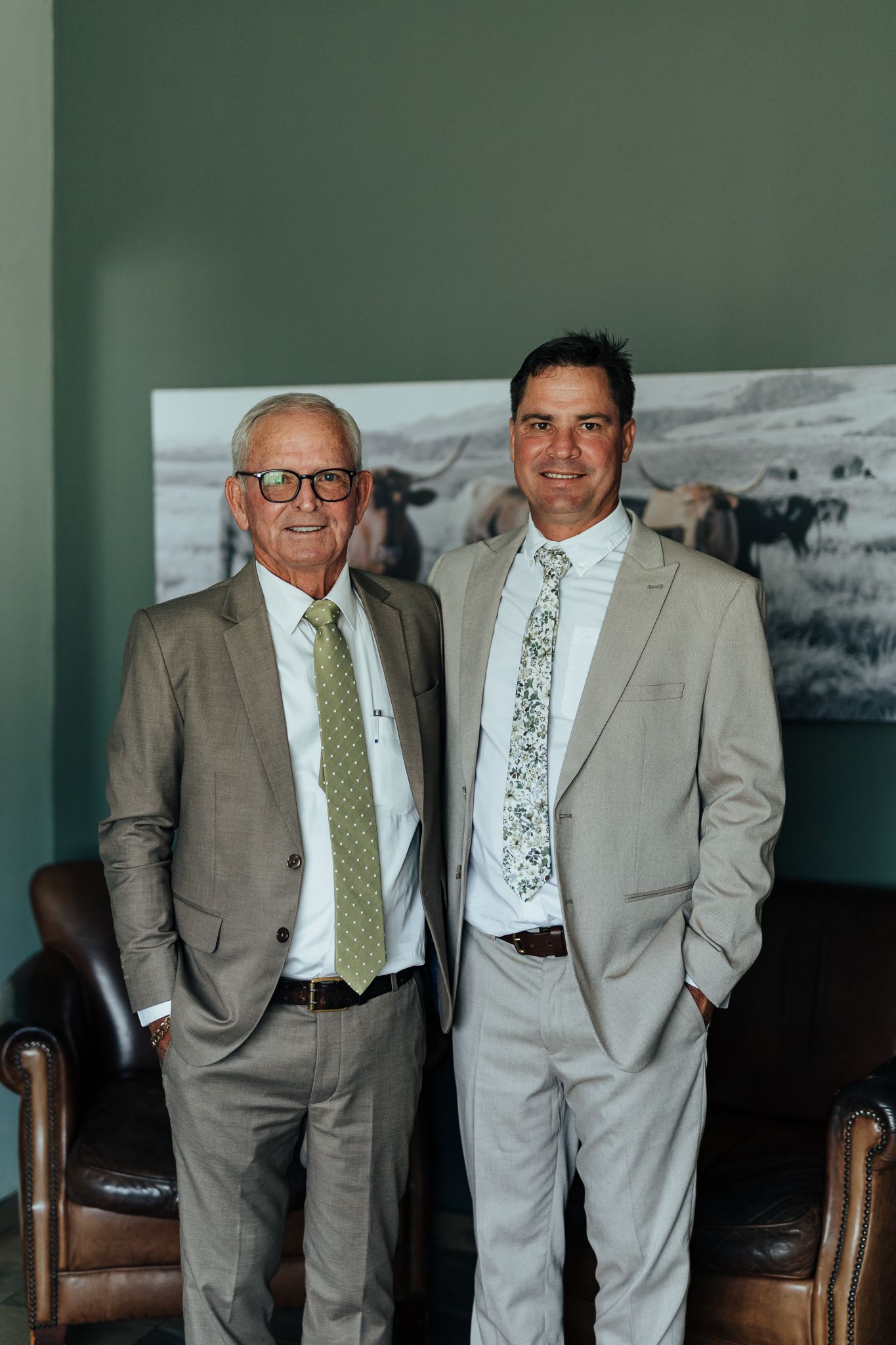 Two men in coordinating gray suits stand together in front of a black and white landscape photo.