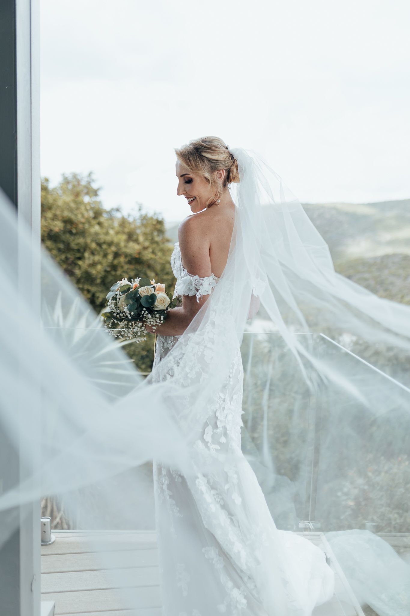 Elegant bride in lace gown with flowing veil poses on balcony with mountain scenery behind.