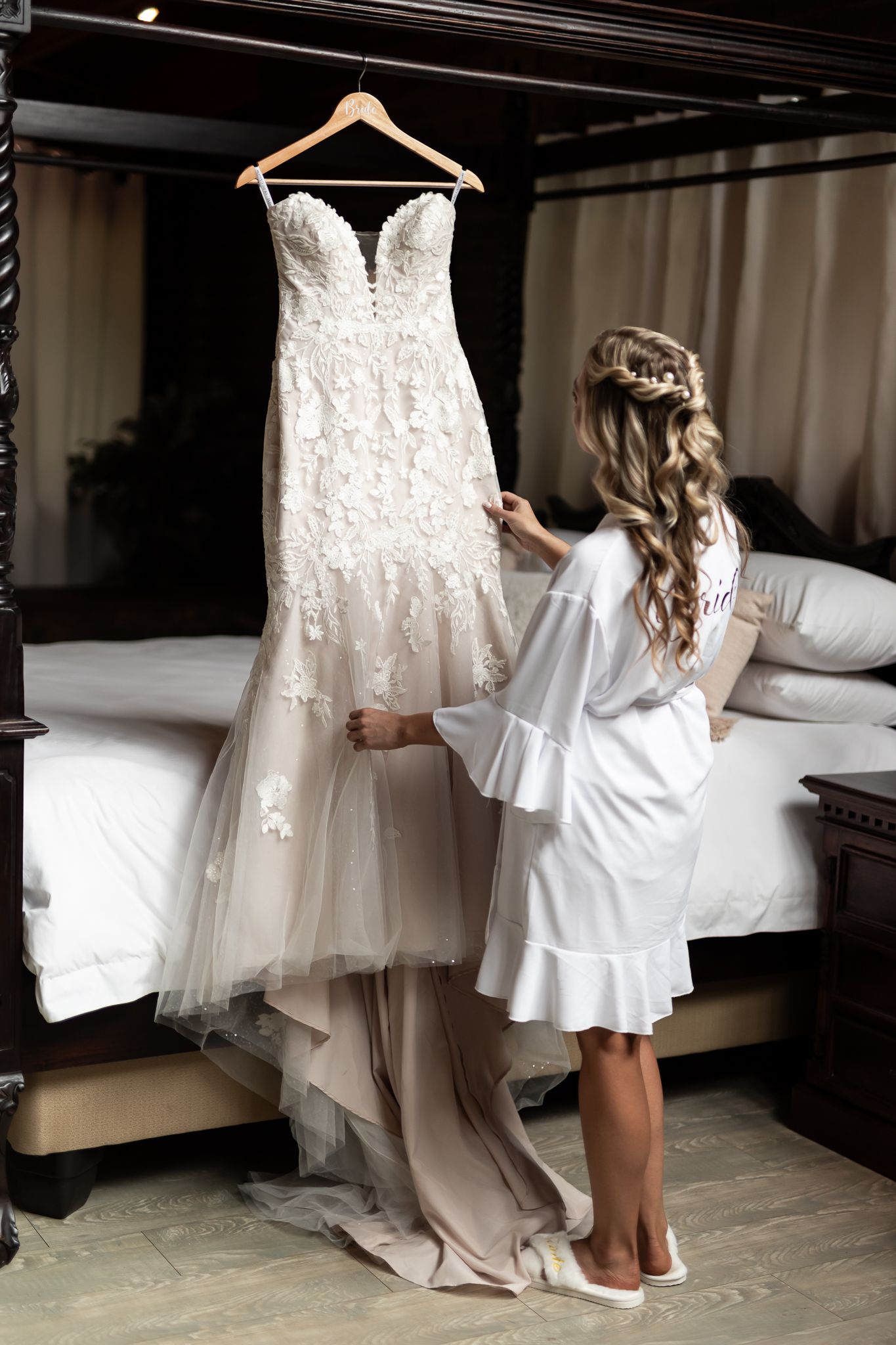 A stunning lace wedding dress hangs while someone in a white robe admires it in a dimly lit room.