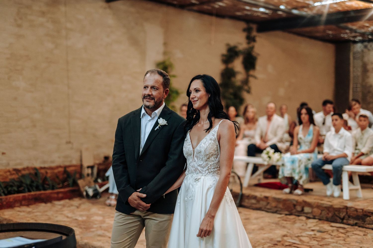 A bride in a white gown and groom in a dark blazer smile while walking down the aisle at an outdoor wedding.