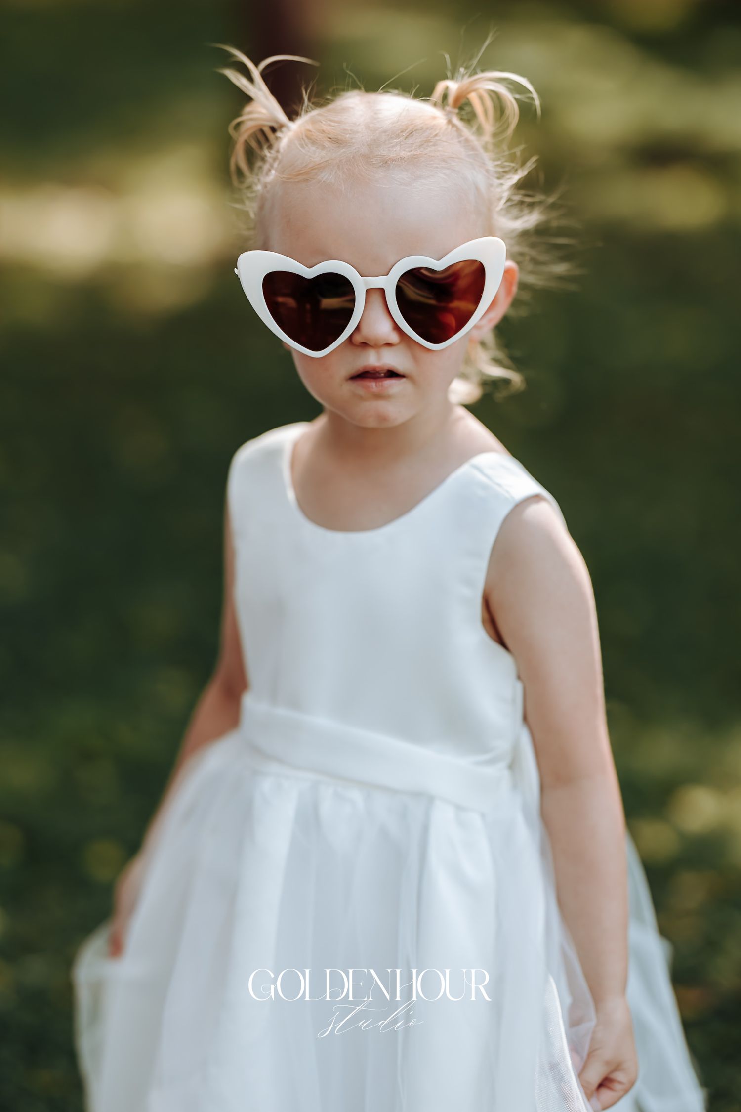 Adorable toddler in white dress wearing heart-shaped sunglasses at an outdoor wedding.