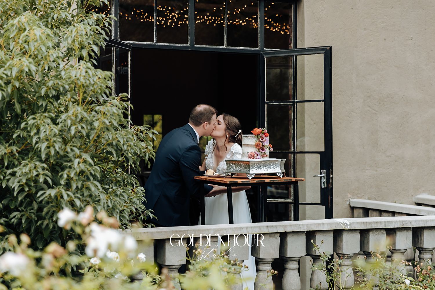 Bride and groom share a romantic kiss while cutting their wedding cake on a balcony.