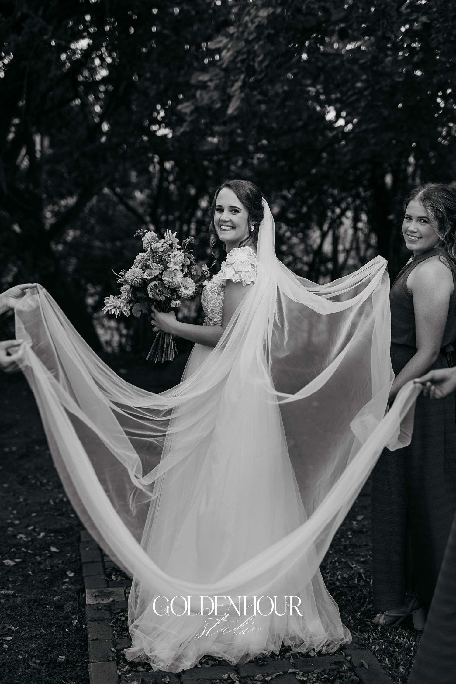 Black and white photo of bride in flowing gown and veil holding bouquet with bridesmaid in garden.