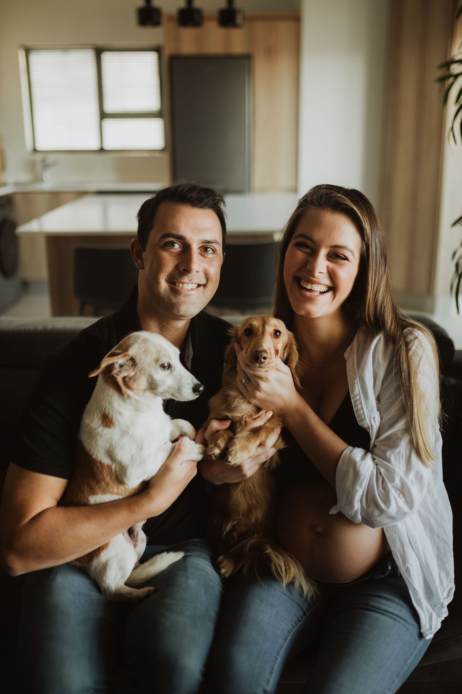 Two women sitting on couch holding white and brown dogs while smiling at camera in home setting.