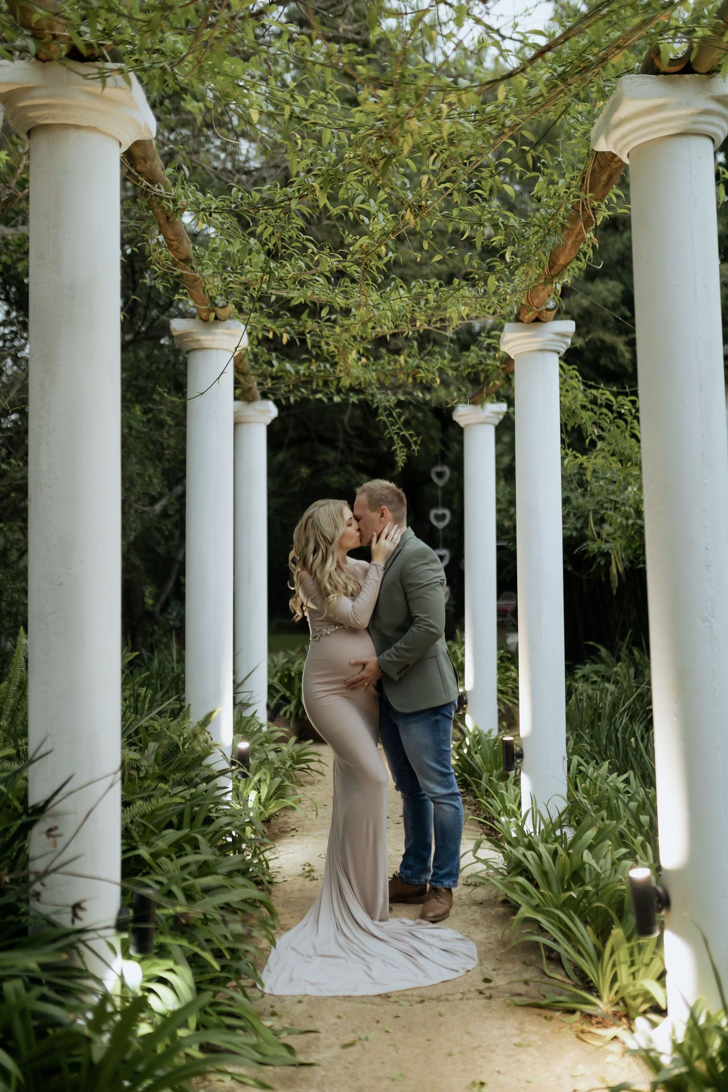 A romantic pregnancy photoshoot under a white columned pergola surrounded by greenery.
