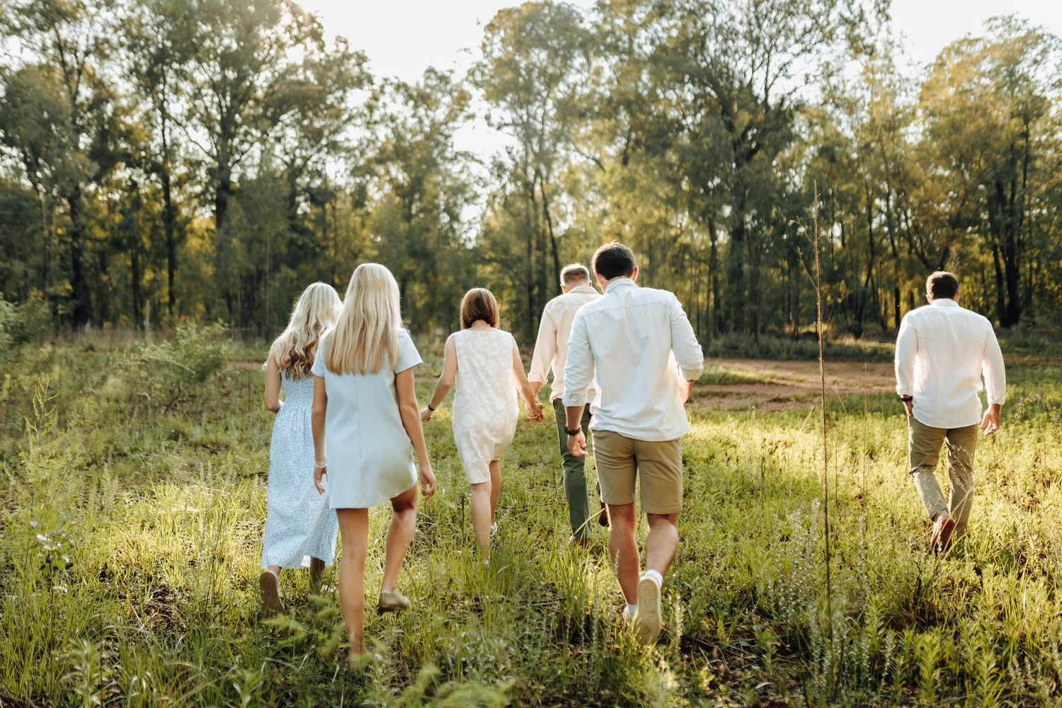 Several people in coordinating white and blue attire walking through a sunlit meadow with tall trees in background.