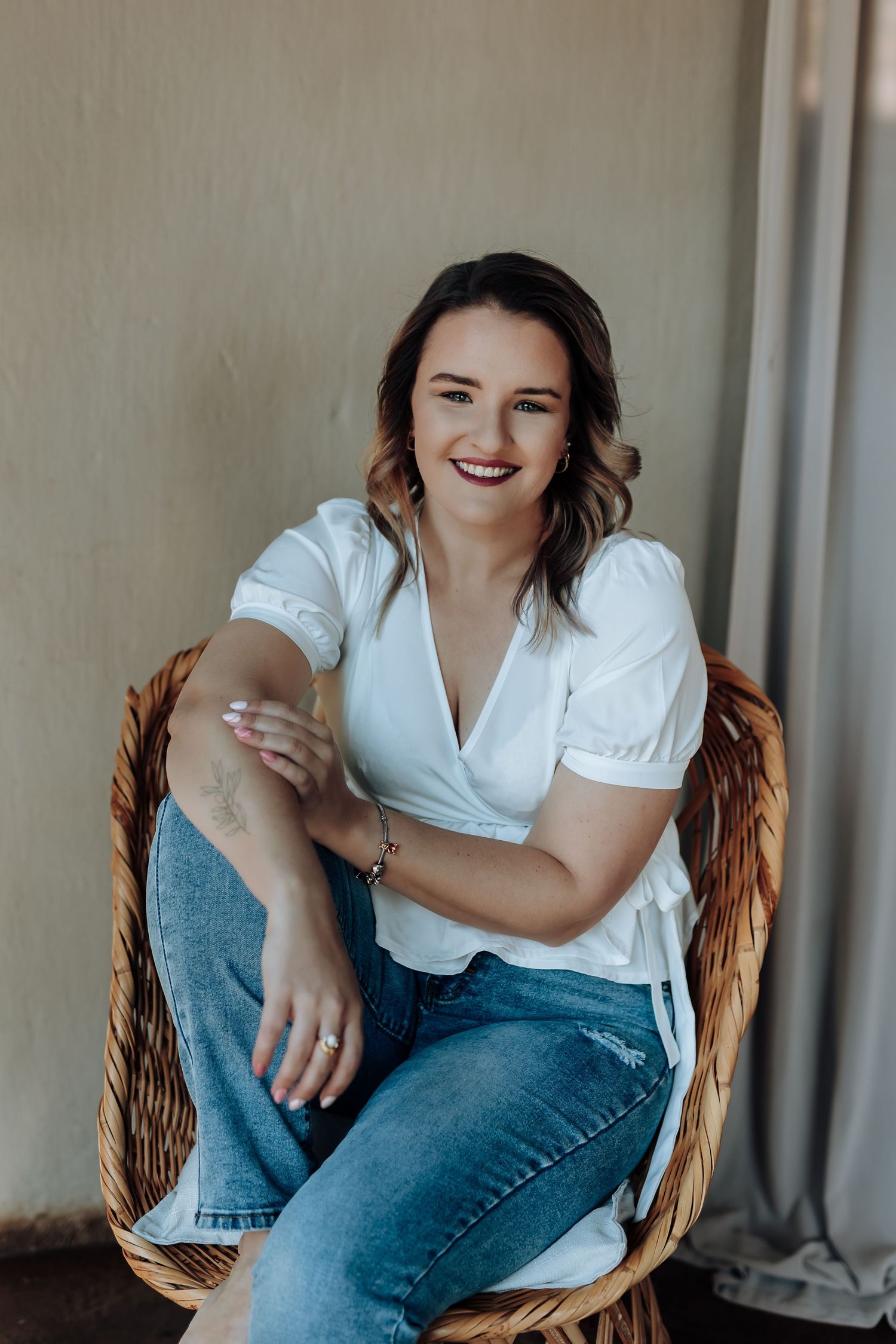 A person in a white shirt and jeans sitting and laughing in a wicker chair during a casual photoshoot.