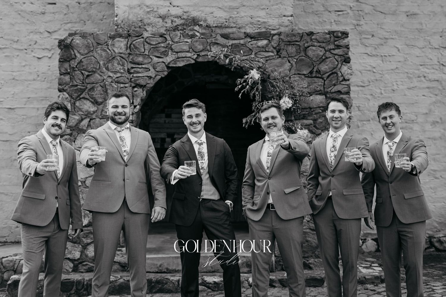 Wedding party in matching gray suits laughing together against stone wall backdrop.