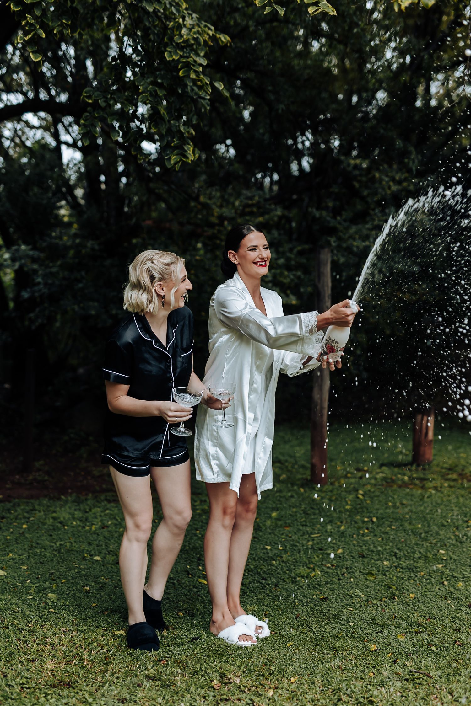 Two women celebrating with champagne outdoors, one spraying the bottle while both laugh joyfully in a garden setting.