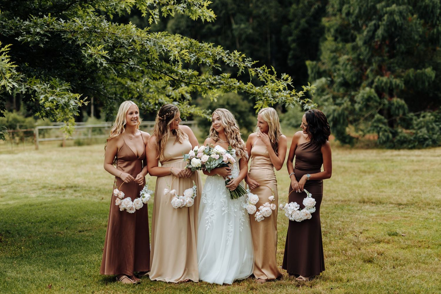 Bridal party wearing earth tone dresses stands together holding bouquets in a sunny outdoor garden setting.
