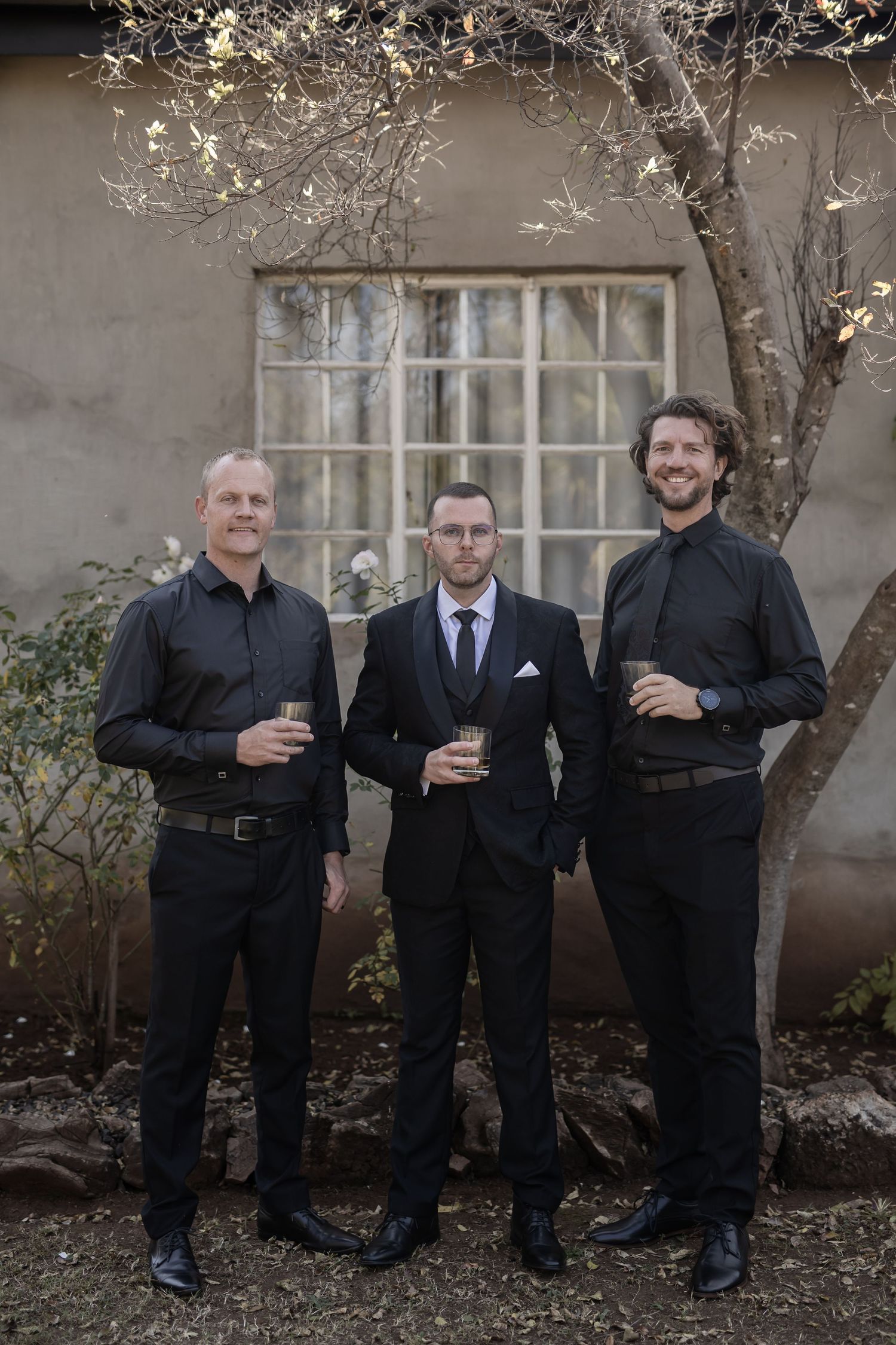 Three well-dressed men in dark attire holding drinks pose together in front of a rustic wall with a window and tree.