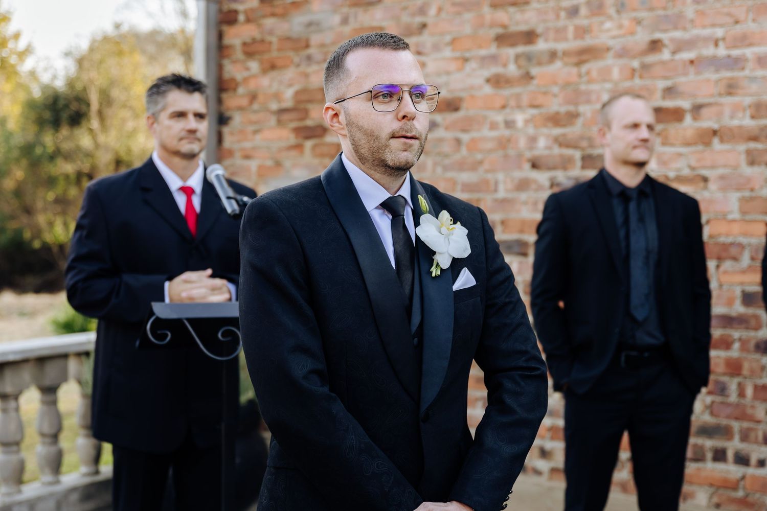 Groomsmen in formal attire stand near brick wall at outdoor wedding venue during daytime celebration.